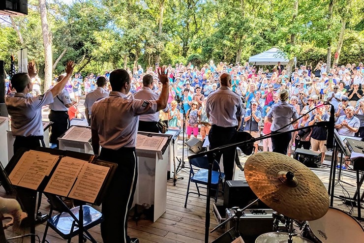 A image of the Heritage Band in blue summer uniform with short sleeves standing on stage in front of a colorful crowd outdoors in front of a green tree backdrop.  Gold percussion instruments are in the foreground.
