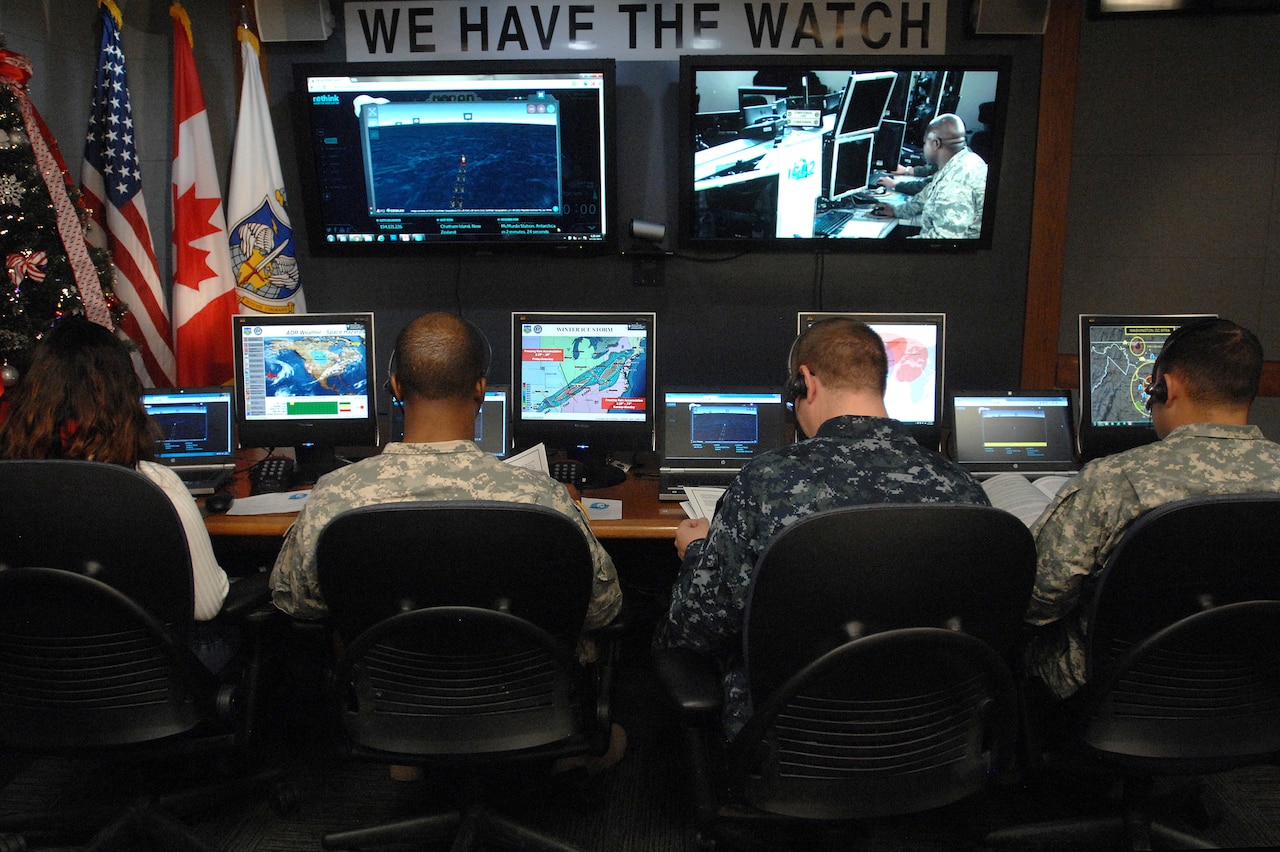 Four people sit facing several monitors. A sign above them reads: "We have the watch."