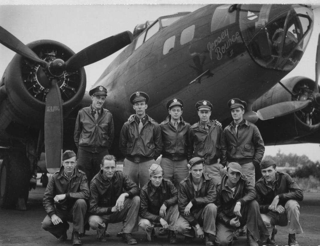 Five men stand and six men take a knee in front of a bomber airplane.
