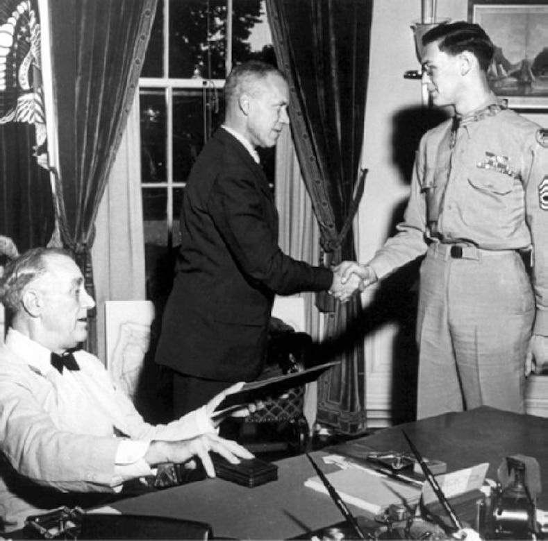 A man sits at a desk while two other men stand behind him, shaking hands.