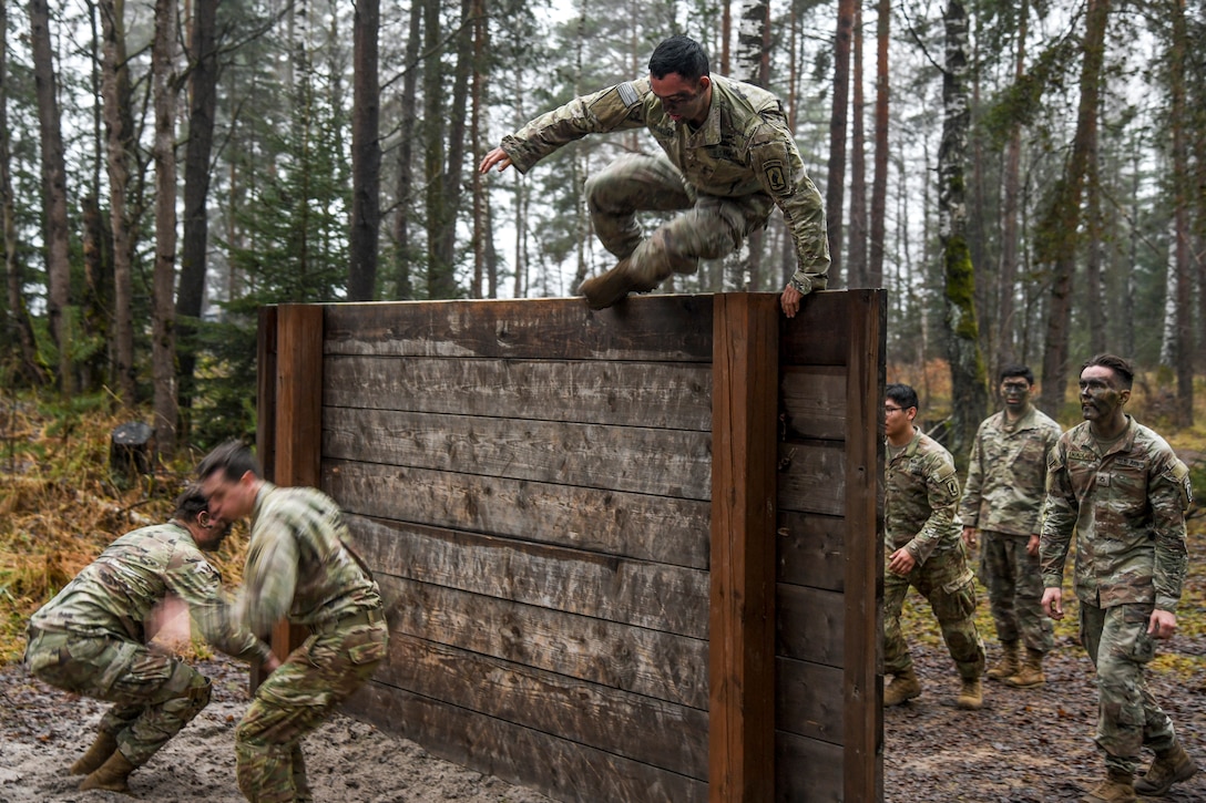A soldier jumps over a wood wall in a forested area as others move on either side of it.