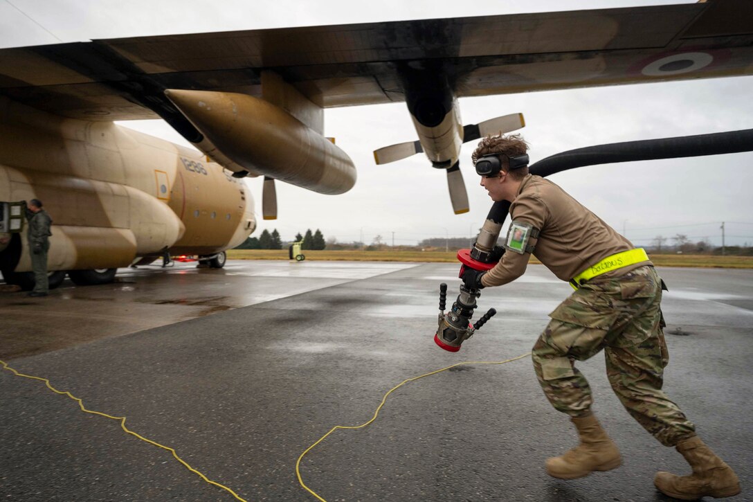 An airman runs with a fuel hose towards an aircraft parked on a tarmac.