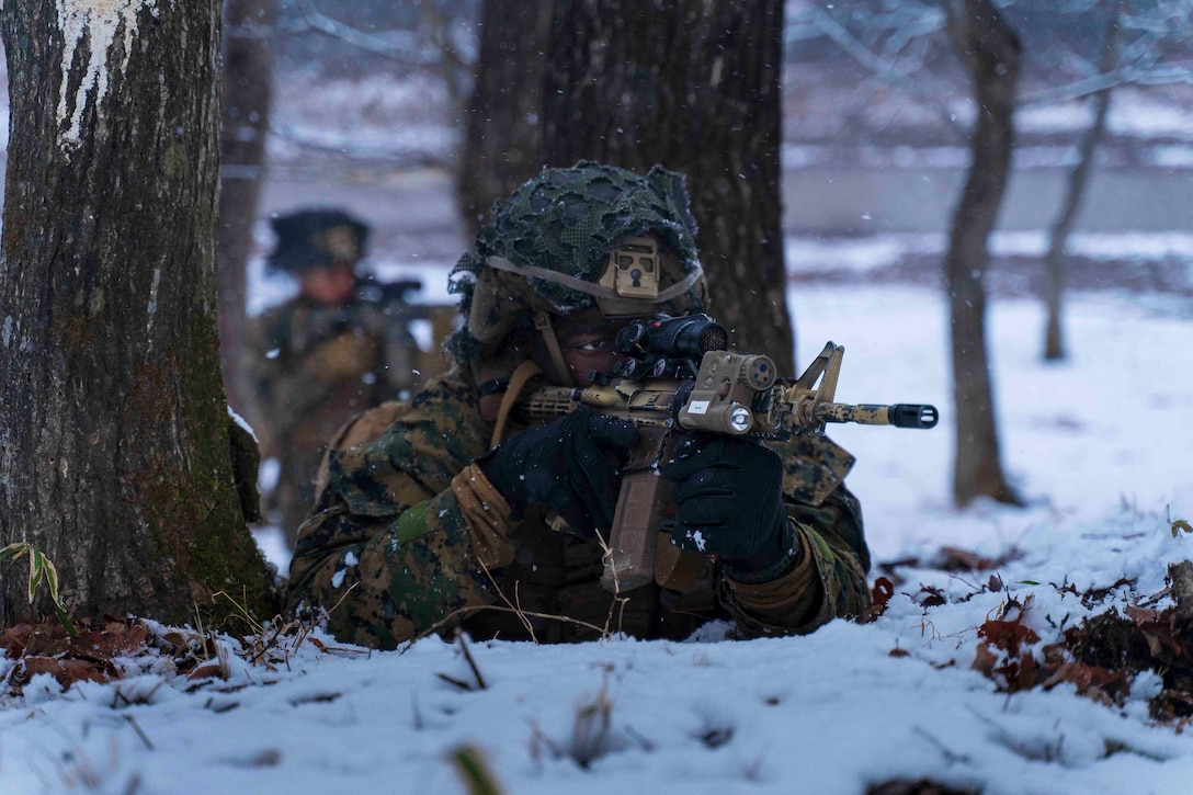 A Marine looks through the scope of a weapon in a wooded area as snow falls.