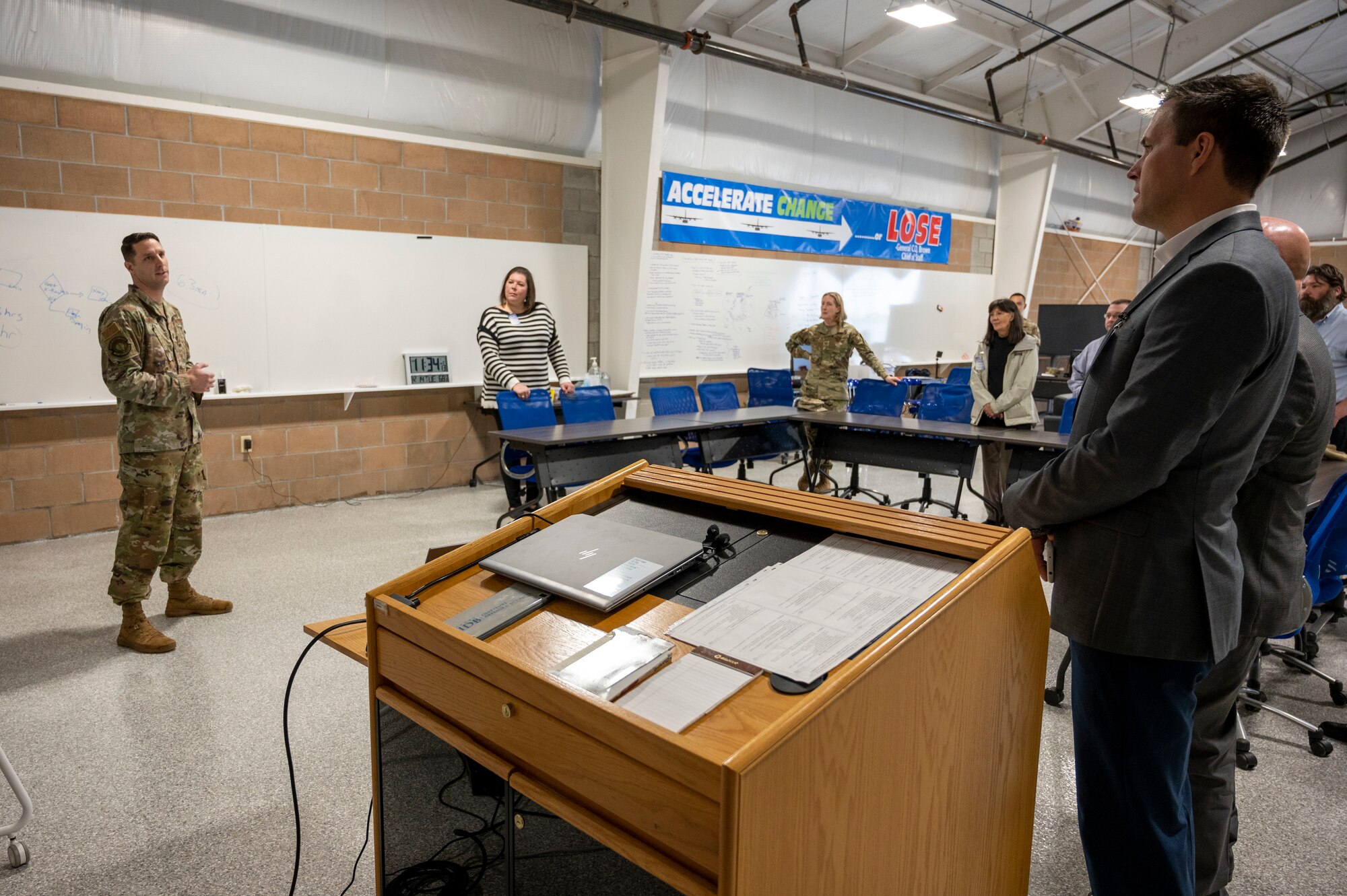Airmen and honorary commanders stand near desks in a classroom and talk.