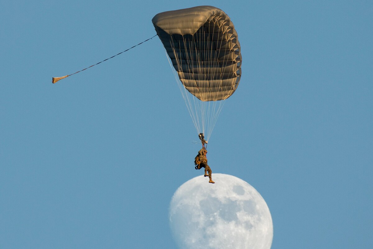 A Marine descends by parachute in blue sky with the moon as backdrop.