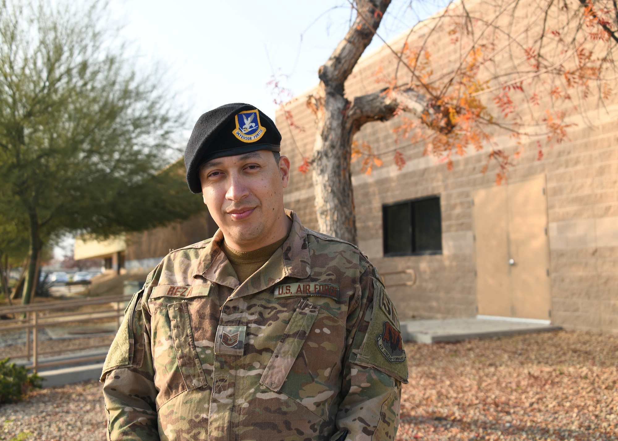A male service member, wearing the operational camouflage pattern and a black beret, facing front, stands outside a building