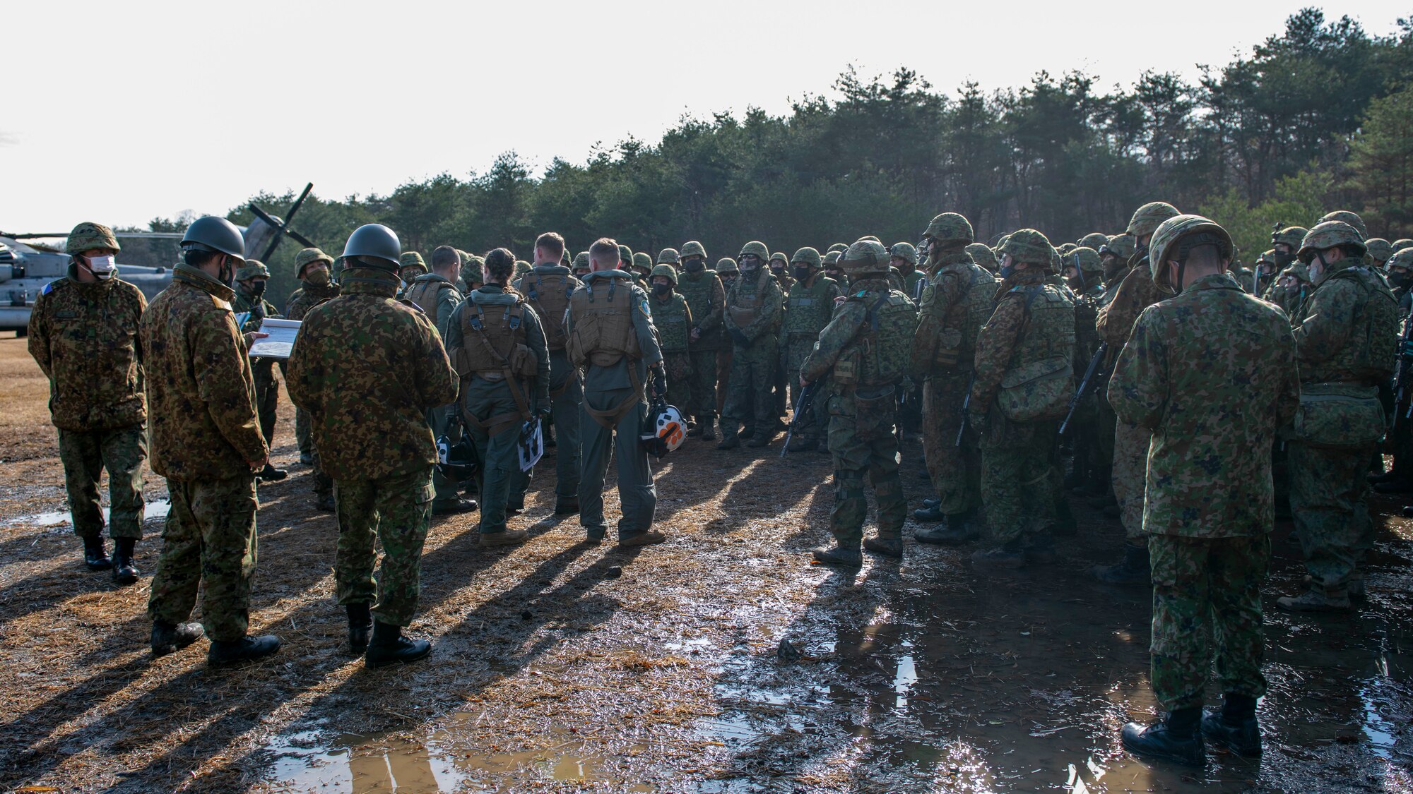 A large group of mixed U.S. and Japanese soldiers stand in a muddy field.