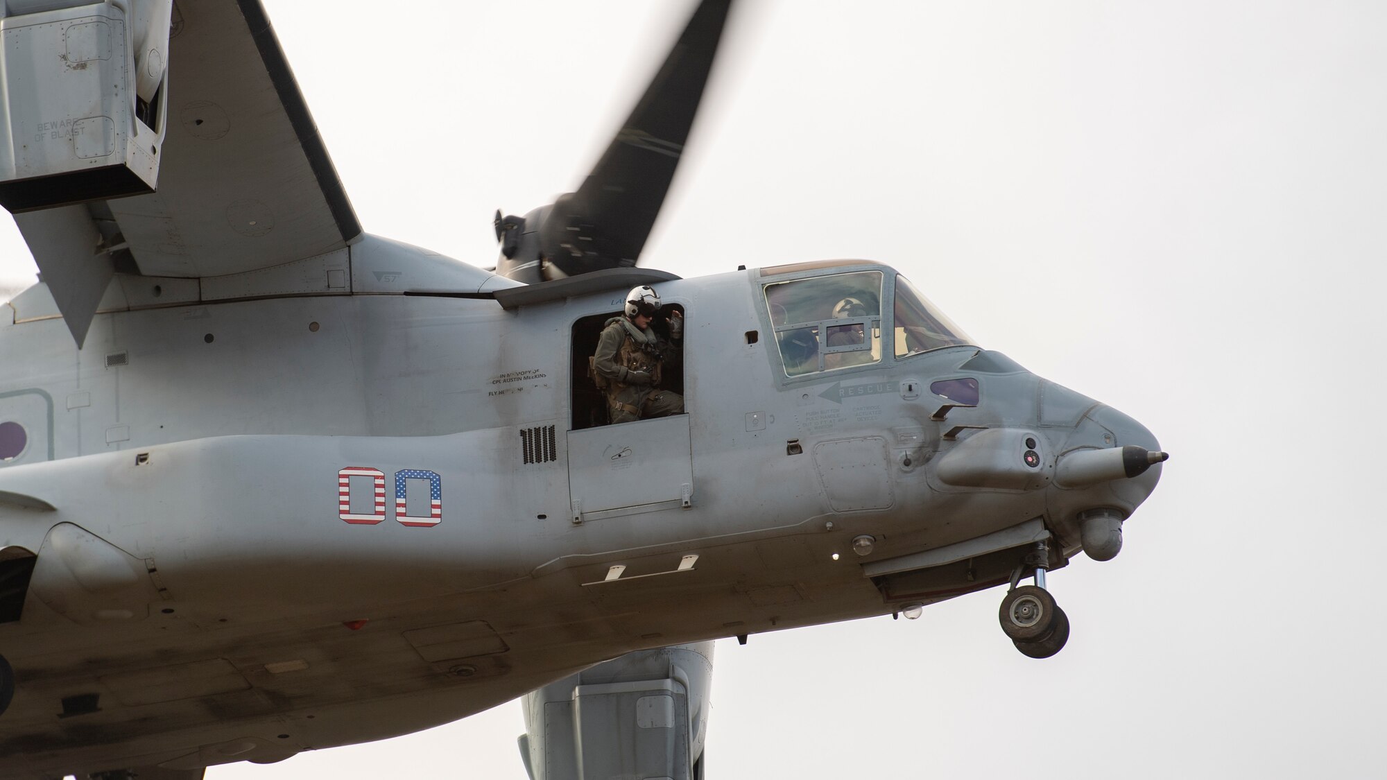 A Marine leans out the door of a hovering aircraft as it comes in for a landing.