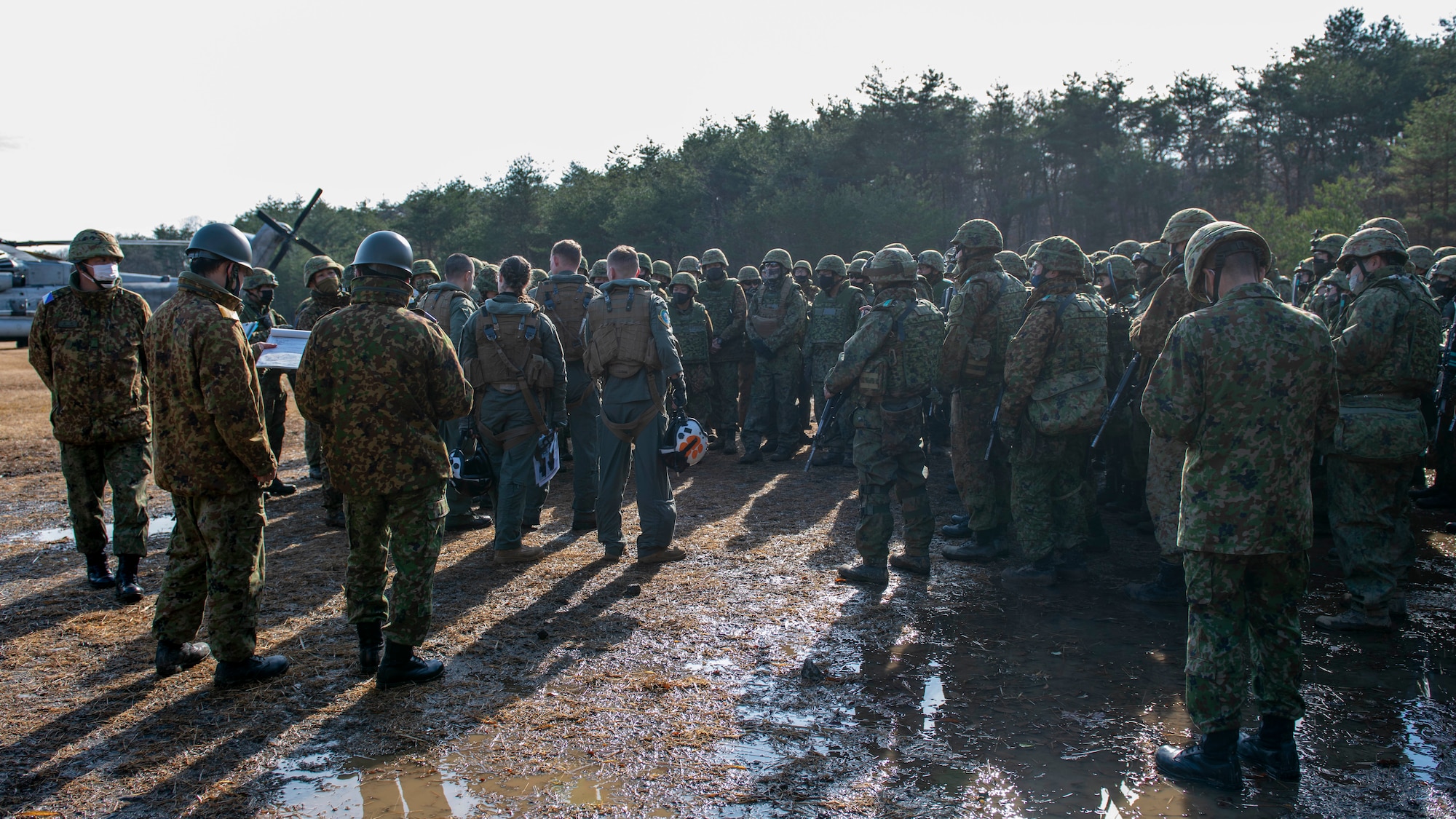 A large group of mixed U.S. and Japanese soldiers stand in a muddy field.