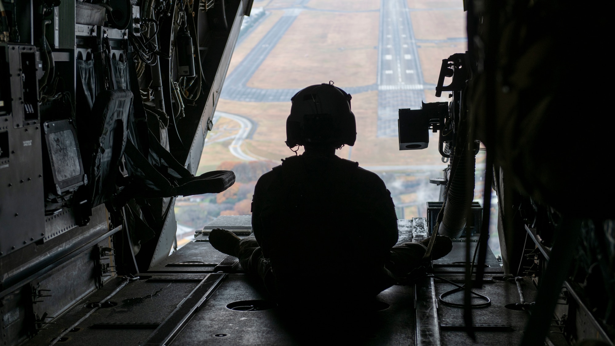 A helicopter flight engineer sits on the edge of an open hatch of an airborne helicopter.