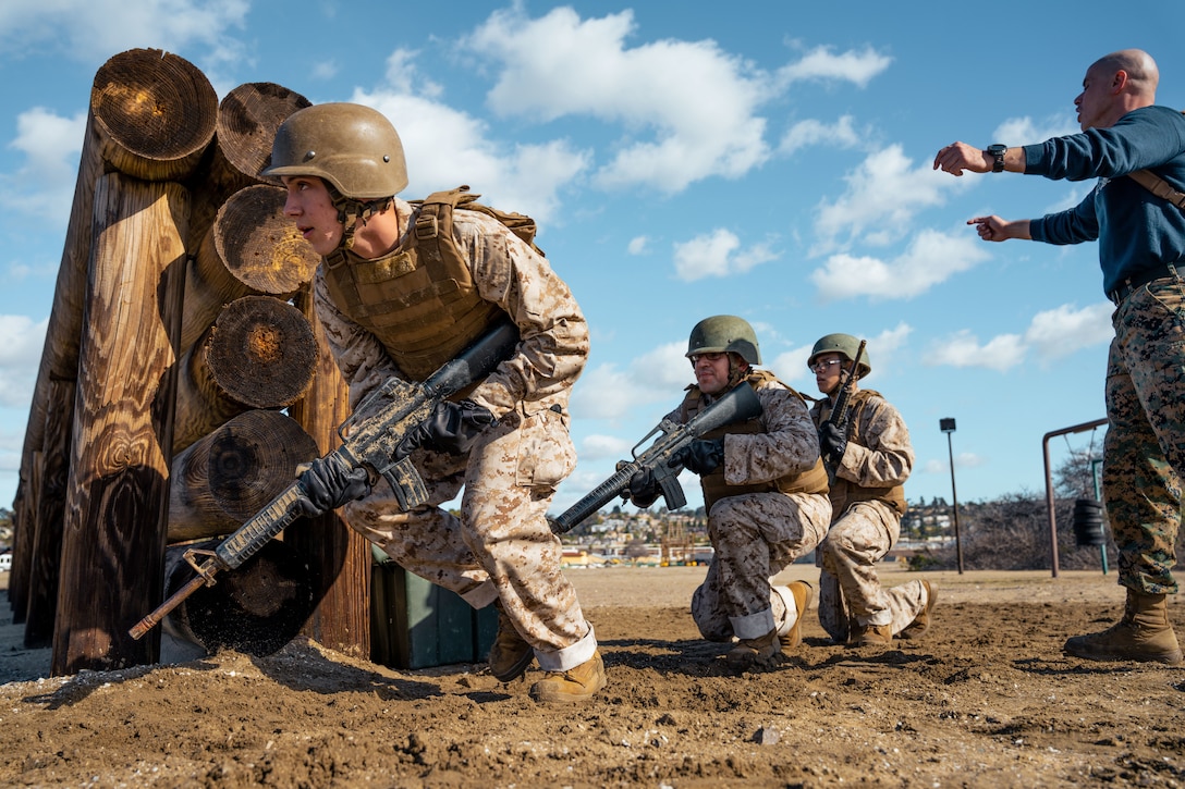 Marine Corps recruits holding weapons kneel around a wooden barrier.