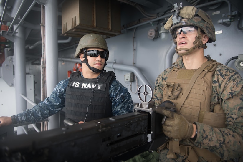 Two service members in combat gear and helmets stand beside each other behind a large gun.