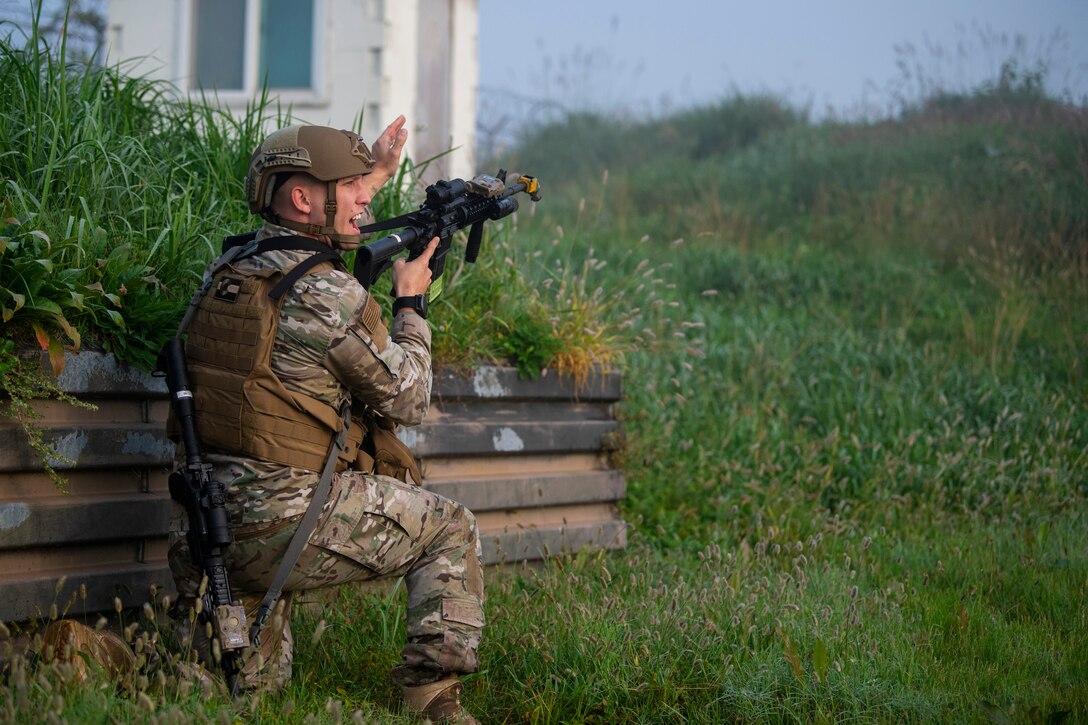 A man with a rifle kneeling beside a wall points into the distance over a rolling grassy hill.