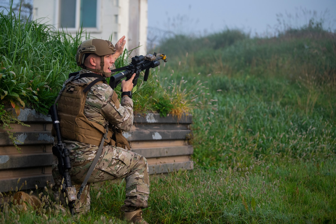A man with a rifle kneeling beside a wall points into the distance over a rolling grassy hill.