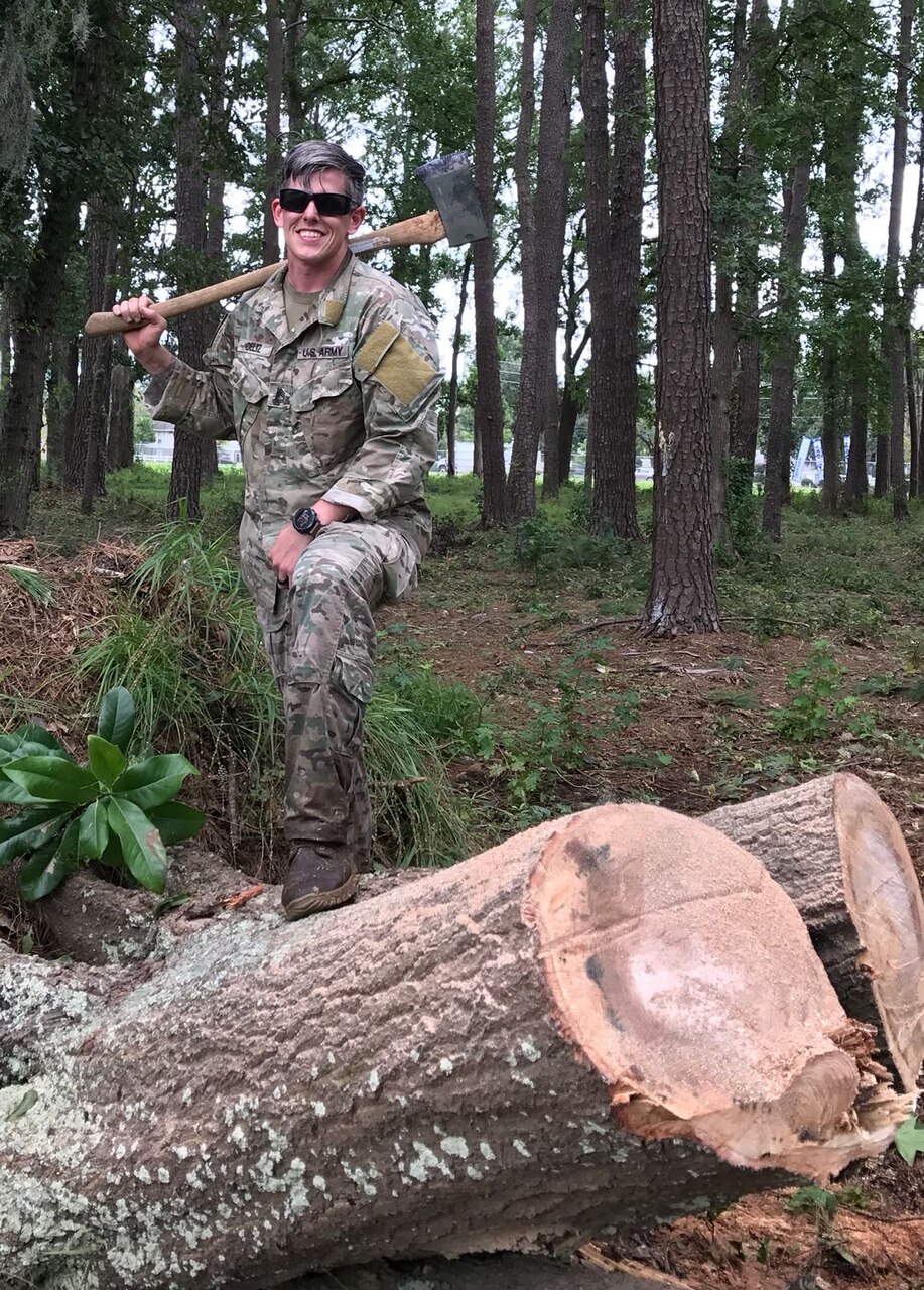 A smiling man holds an ax over his shoulder as he kneels on the ground; tall trees are in the background.