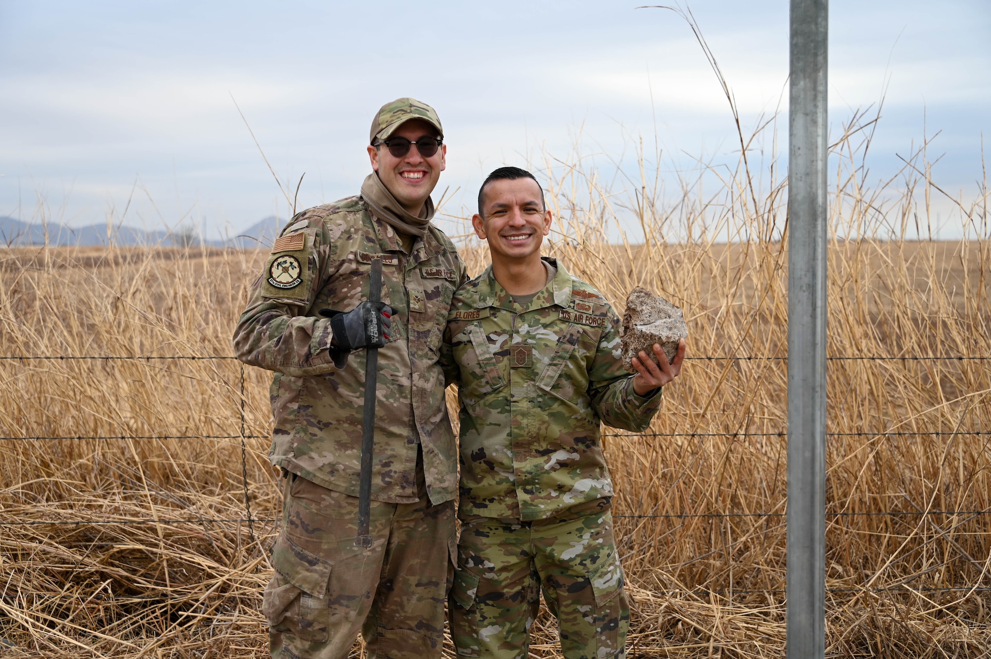 U.S. Air Force Senior Airman James Gonzales, 97th Civil Engineering Squadron equipment operator, and Chief Master Sergeant Ceasar Flores, 97th Air Mobility Wing command chief, pose for a photo at Altus Air Force Base, Oklahoma, Dec. 17, 2021. Flores helped Gonzales remove a pin from a fence with the rock he is holding. (U.S. Air Force photo by Airman 1st Class Kayla Christenson)