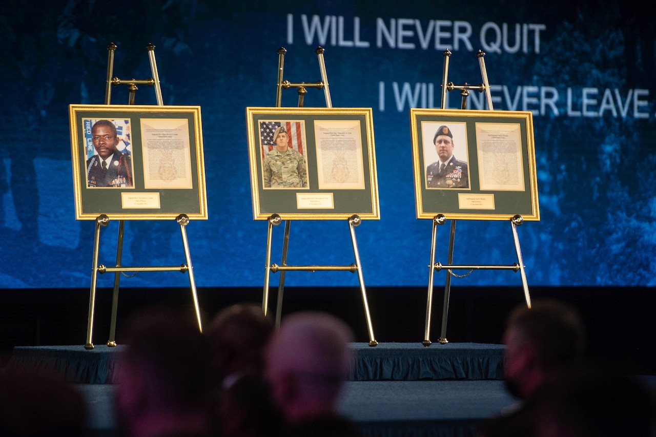 Photos of three soldiers are displayed on stands on a stage in front of a seated audience.