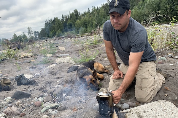 Aan holds a coffee pot over some coals as a dog watches; tall trees are in the background.