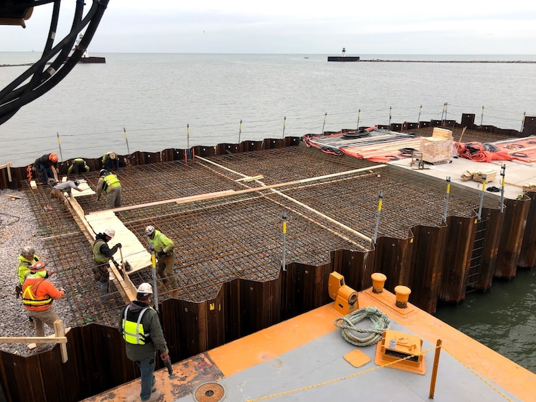 Construction workers place rebar and concrete inside metal sheetpile walls in a harbor.