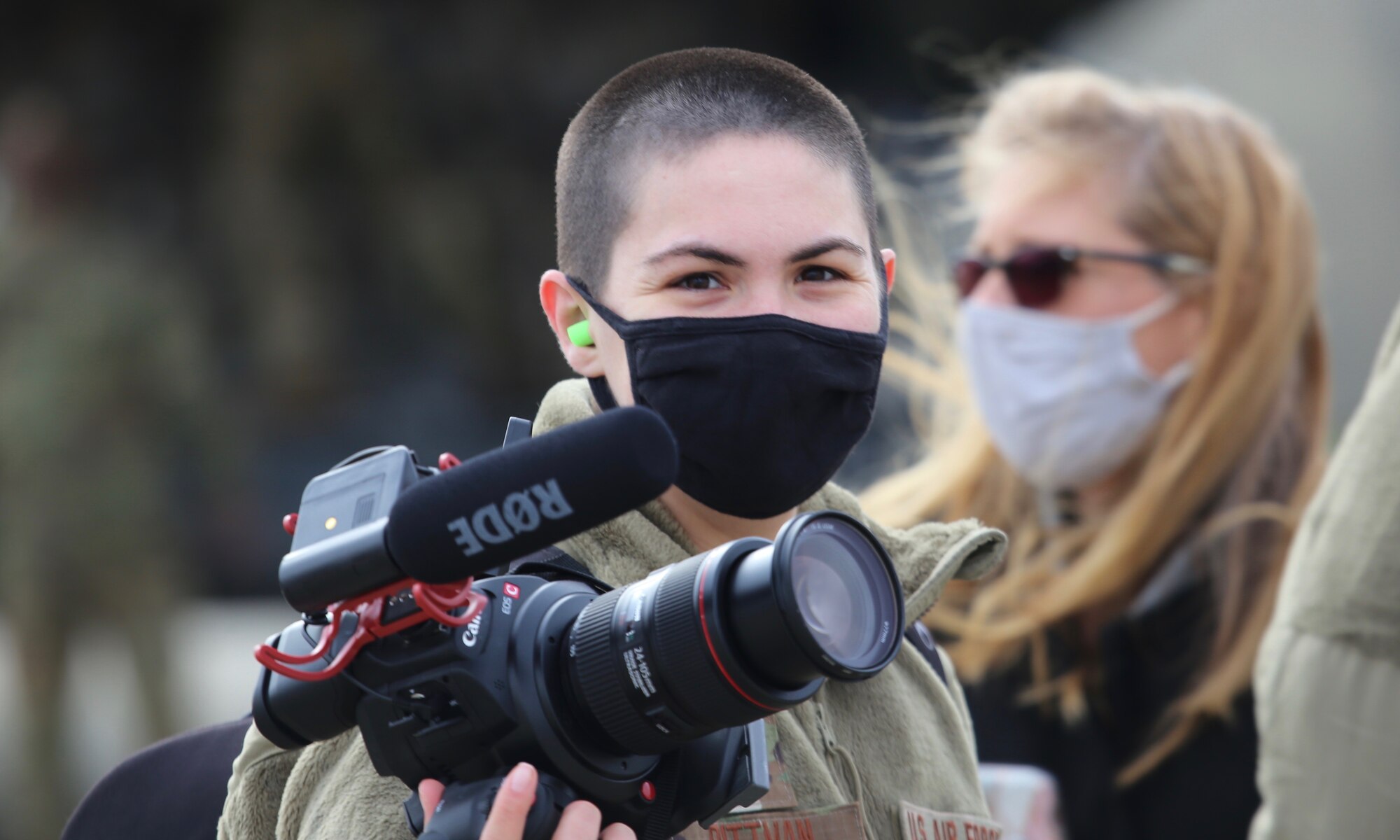 An Airman looks at the camera, while holding a camera. Woman in background.