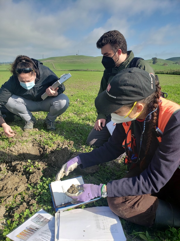 Two white females and one white male look at dirt in a green field and compare it to information in a white book.