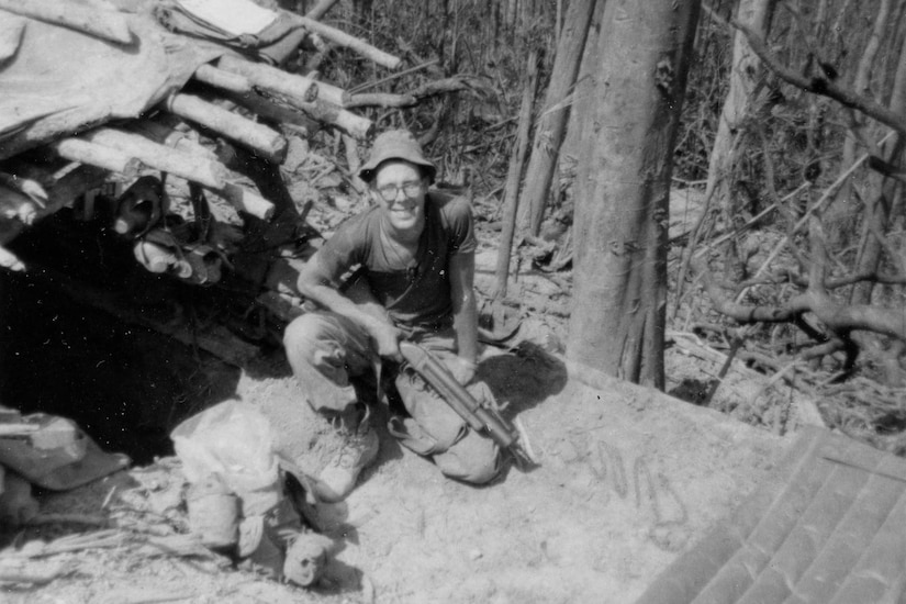 Marine squats on the ground and looks up for a photo; behind him, trees are stripped of their leaves.