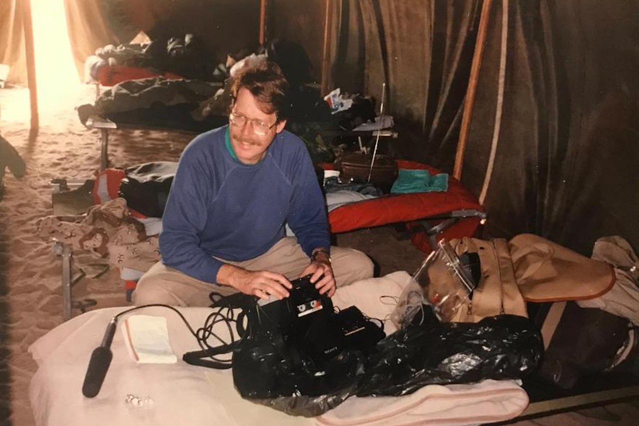 A man squats next to a cot that holds equipment.
