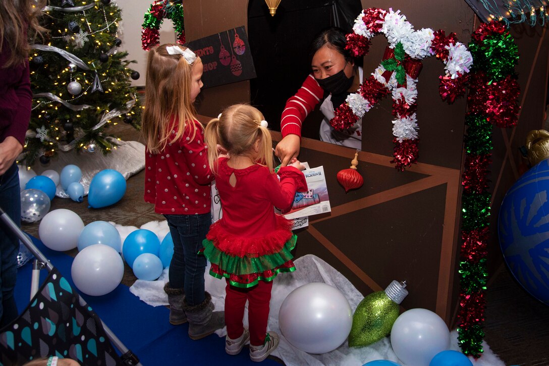 One of Santa's helpers hands a gift to one of two children surrounded by Christmas decorations.