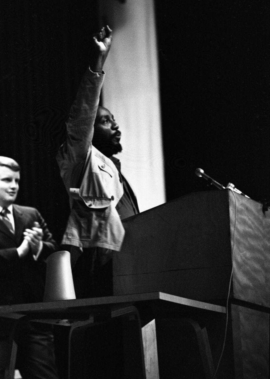 A man holds his fist in the air at a podium as another man applauds behind him.