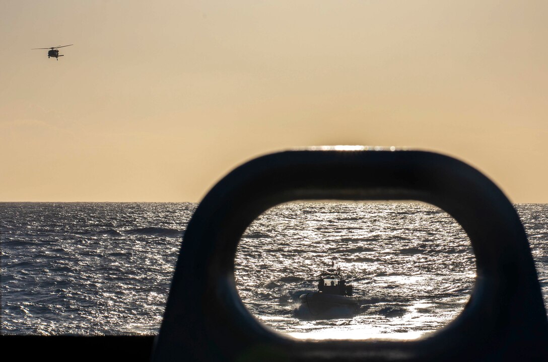 A boat in the ocean is seen from a ship, while a helicopter flies overhead.