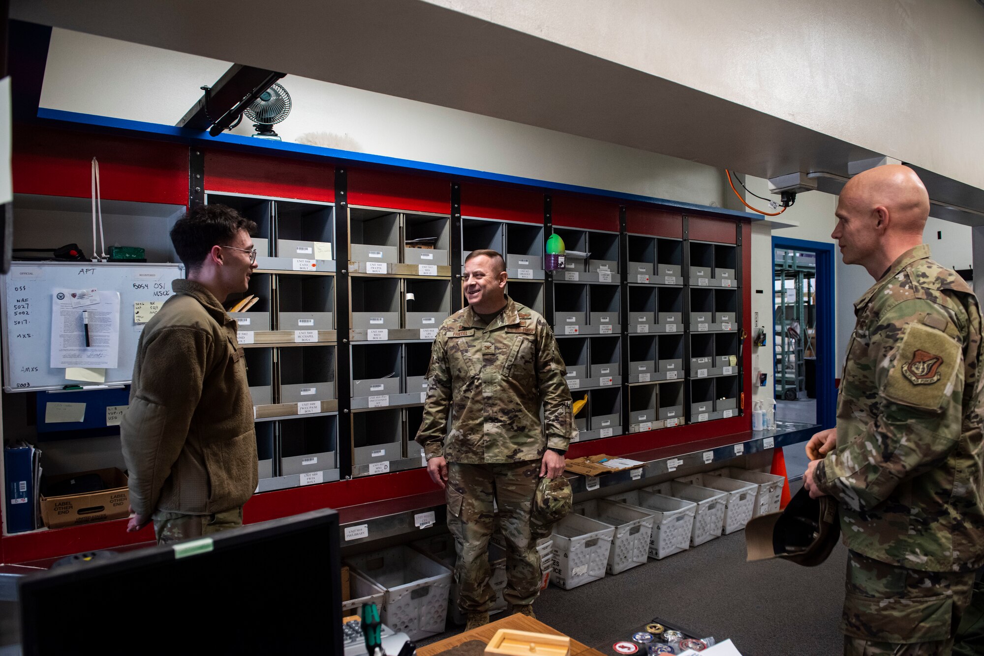 A group of military members in uniform talk next to shelves of mail.
