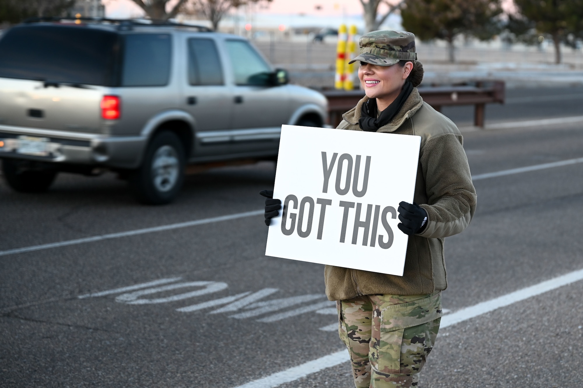 A female servicemember holds a sign with a positive saying at an installation gate.