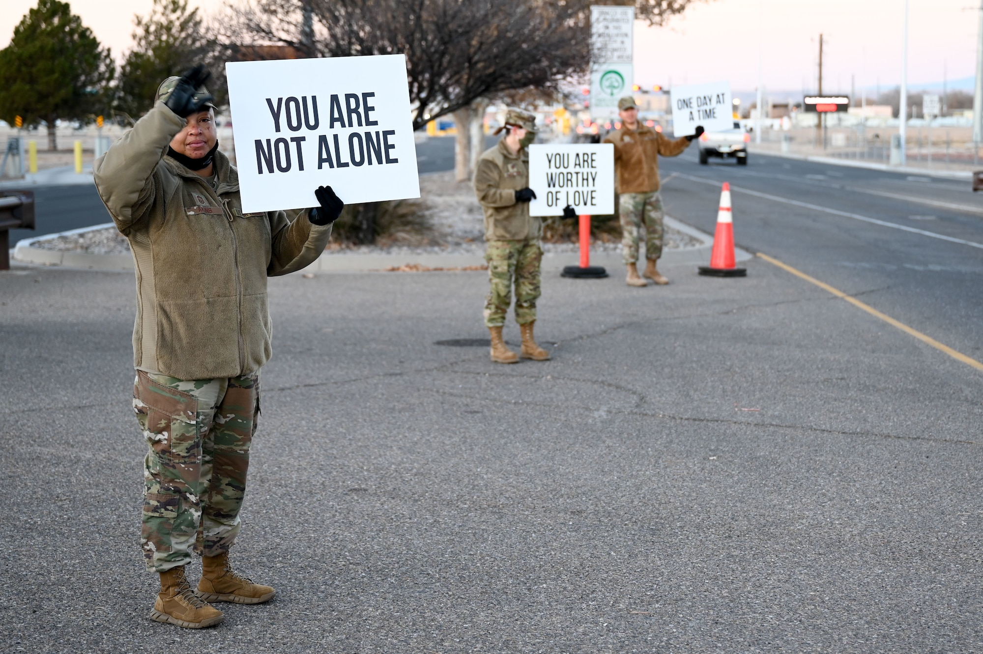 Three servicemembers hold signs with positive sayings at an installation gate.