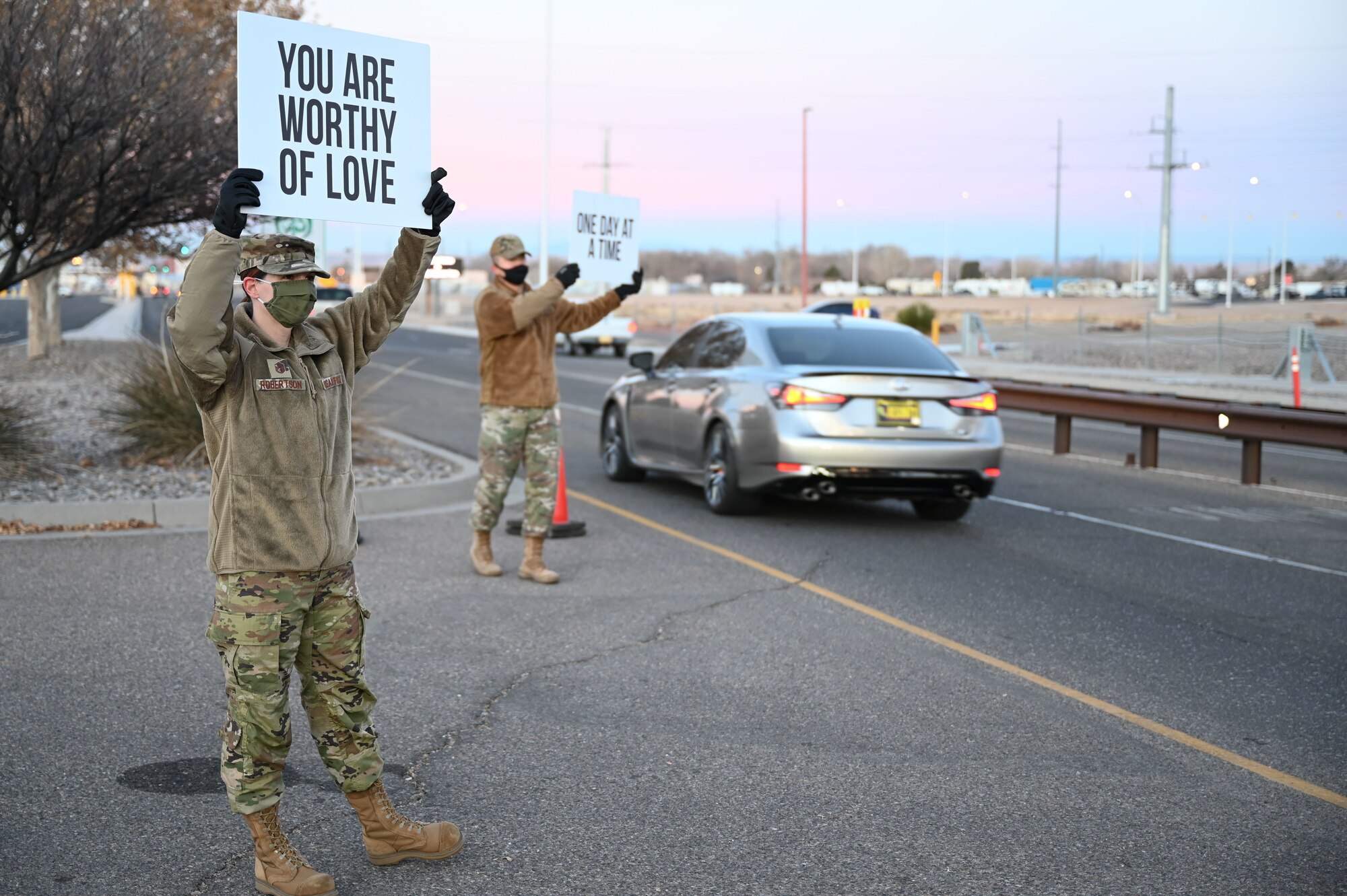 Two servicemembers hold up signs with positive sayings at an installation gate.