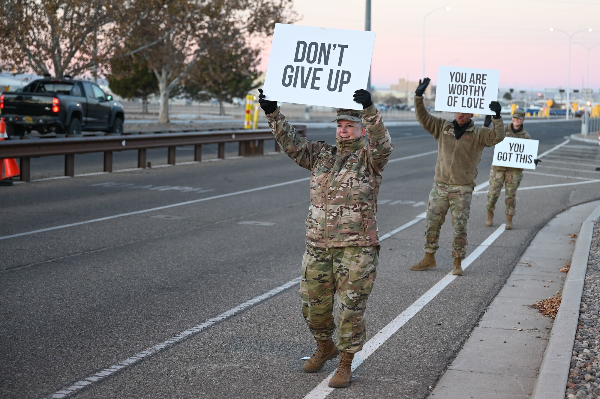 Three servicemembers hold signs with positive sayings at an installation gate.