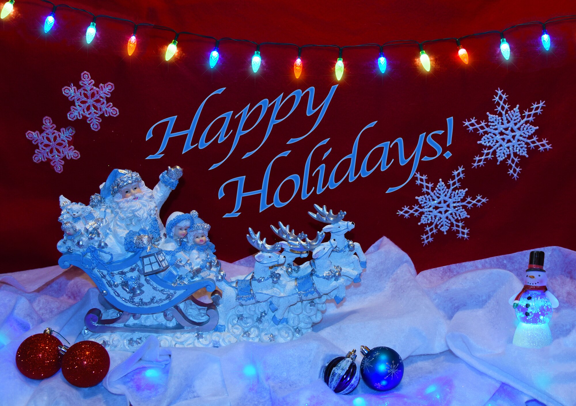 Santa in sleigh pulled by reindeer, tree ornaments in red and blue, snowman with snowglobe on blanket of snow and lights.