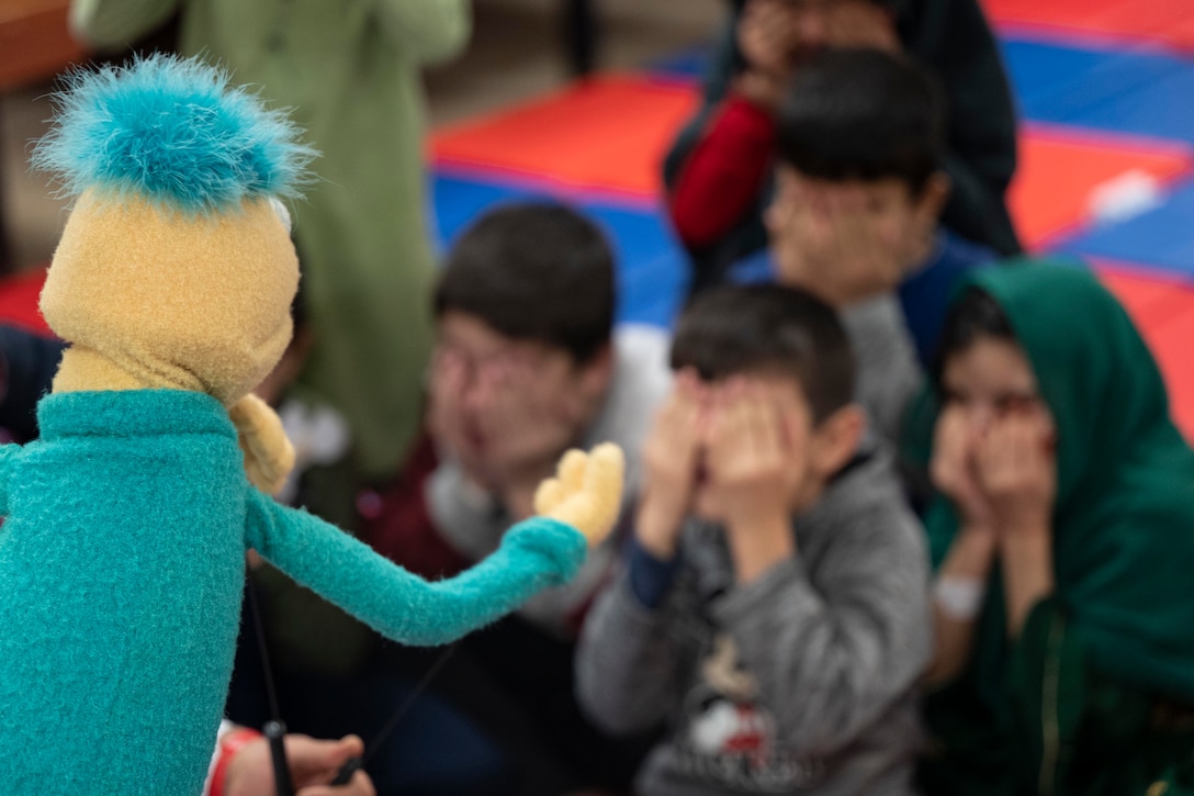 Afghan children cover their eyes while a Muppet performs in the foreground.