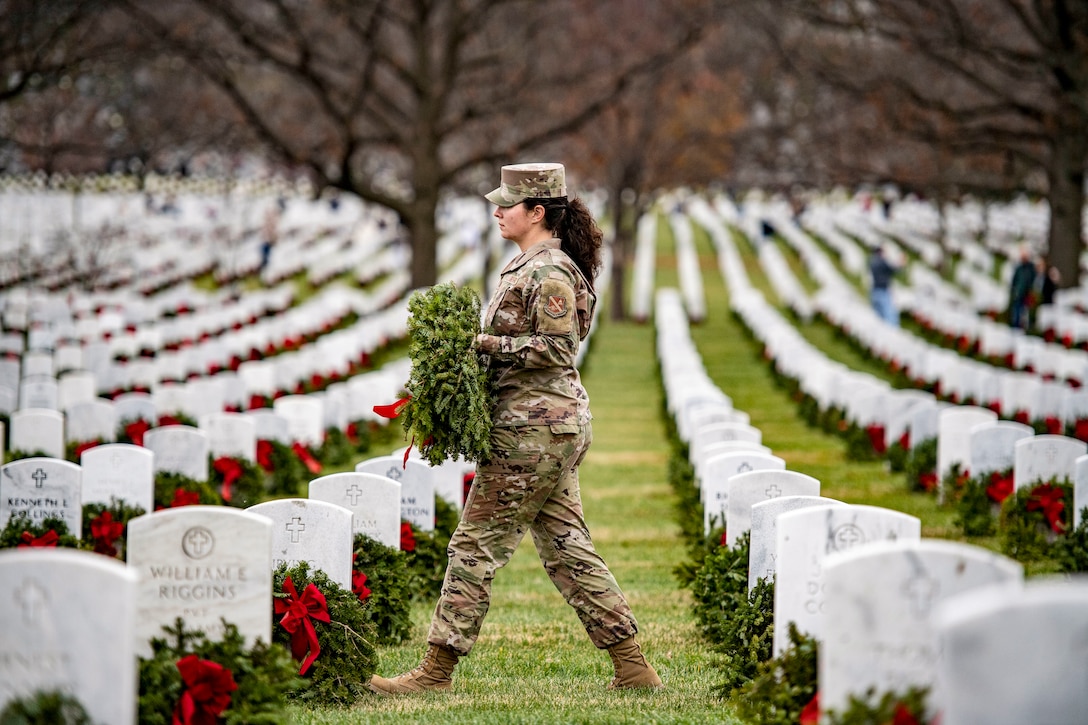 A service member walks with a wreath through a cemetery with wreaths adorning the gravesites.