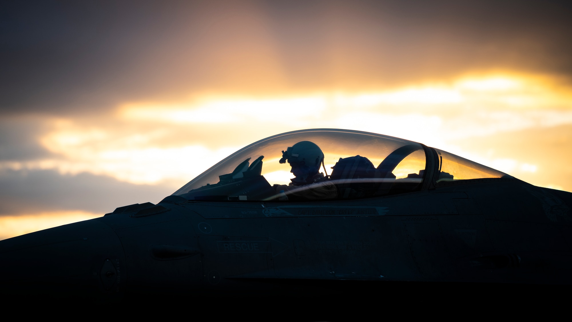 A pilot sits in the cockpit of an F-16C Fighting Falcon in front of a sunset.