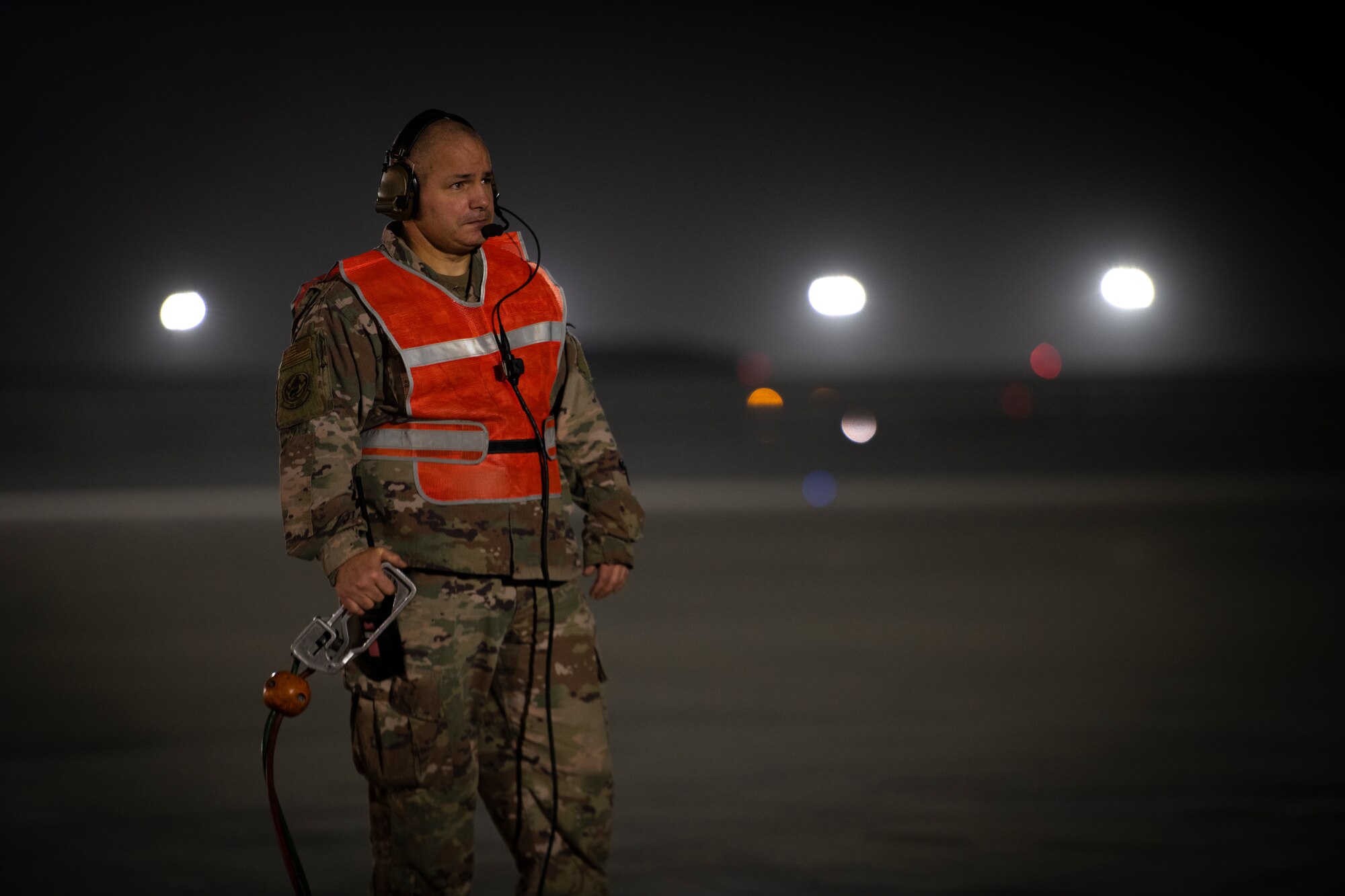 Military member watches plane be refueled while wearing a reflective safety vest.