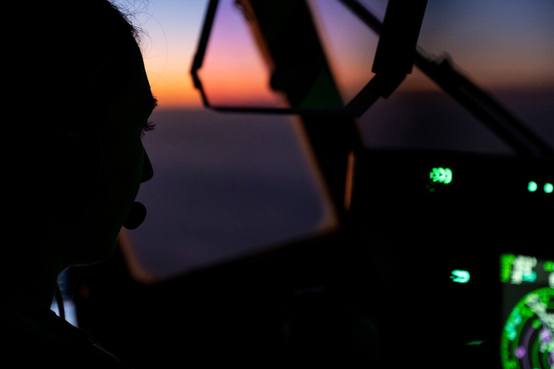 A pilot sitting in the cockpit flies under a sunlit sky as seen in silhouette illuminated by green light.