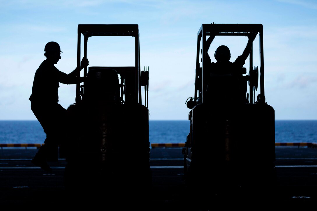 Two sailors shown in silhouette operate two forklifts.