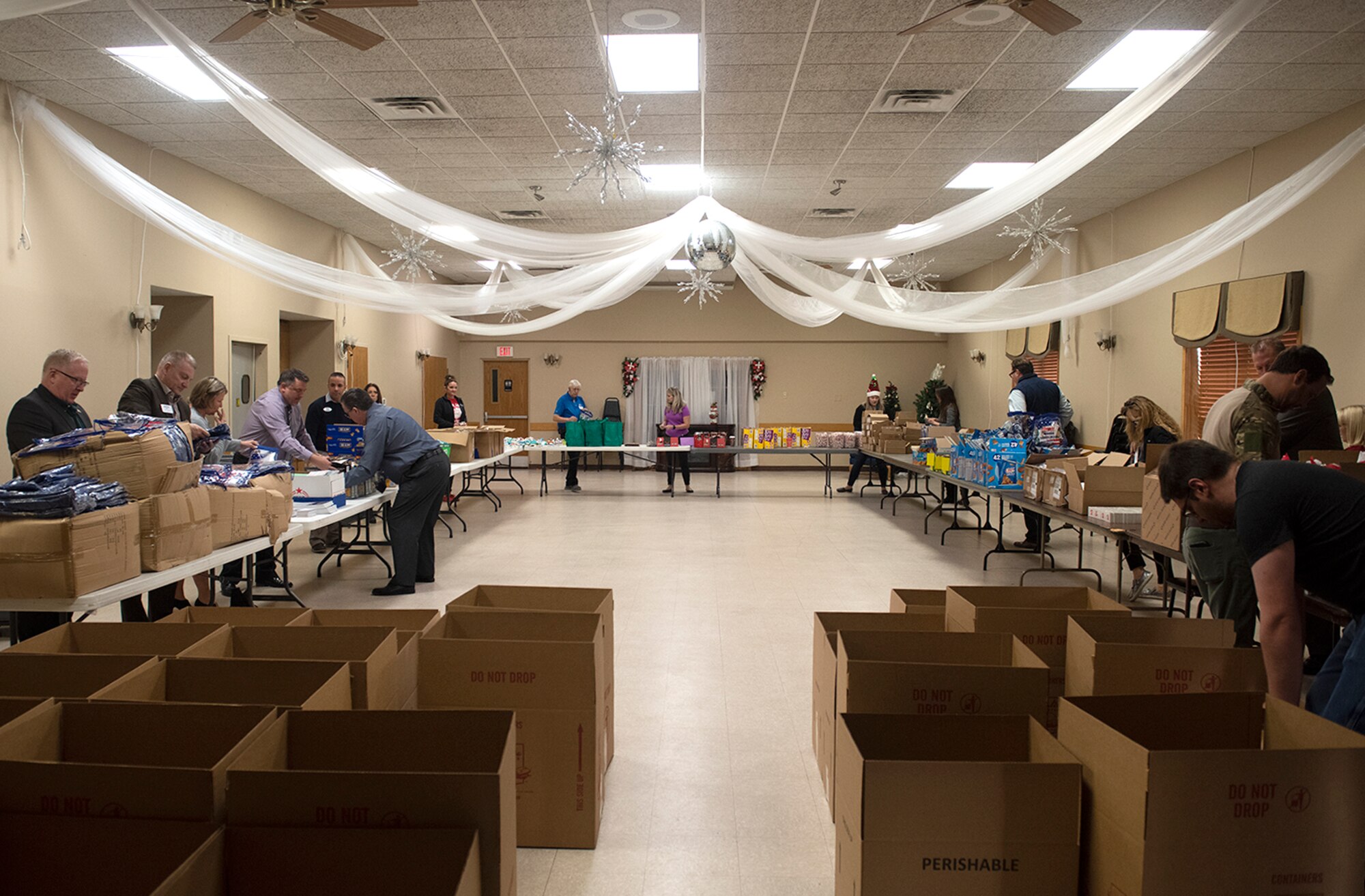 People fill goodie bags from boxes on tables in a large room.