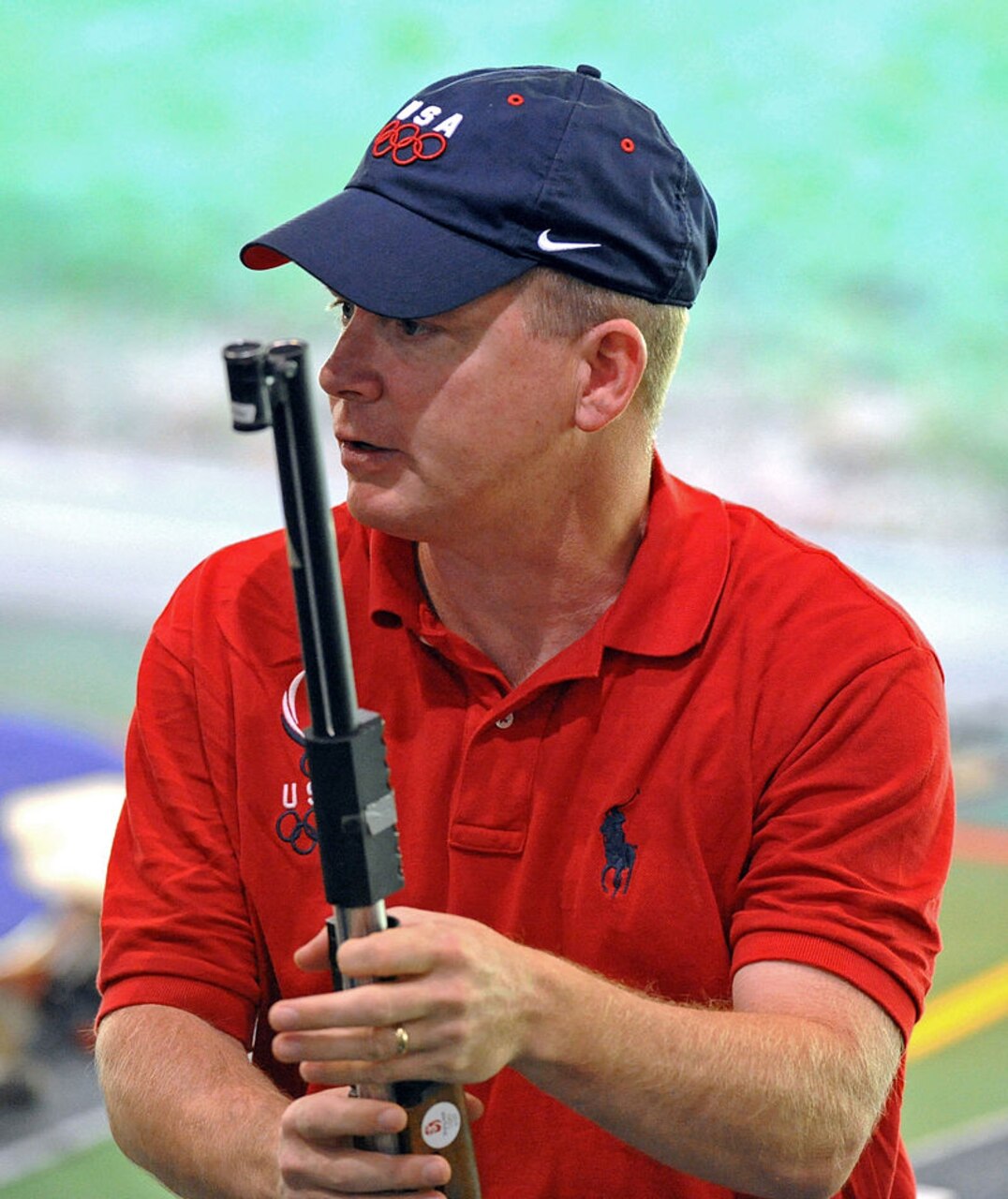 Shooter in USA Olympic hat and shirt looks downrange while holding his rifle.