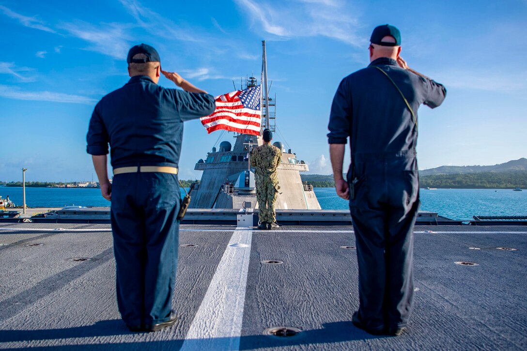 Two sailors salute as another sailor raises an American flag.