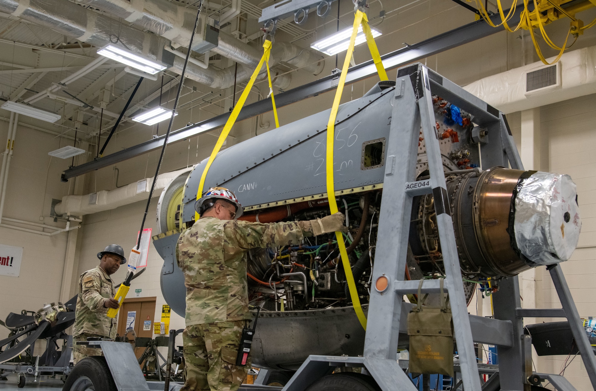 An engine in the middle, a man to the left operates a hoist, while a man in front of the engine hold on to two harnesses cradling the bottom