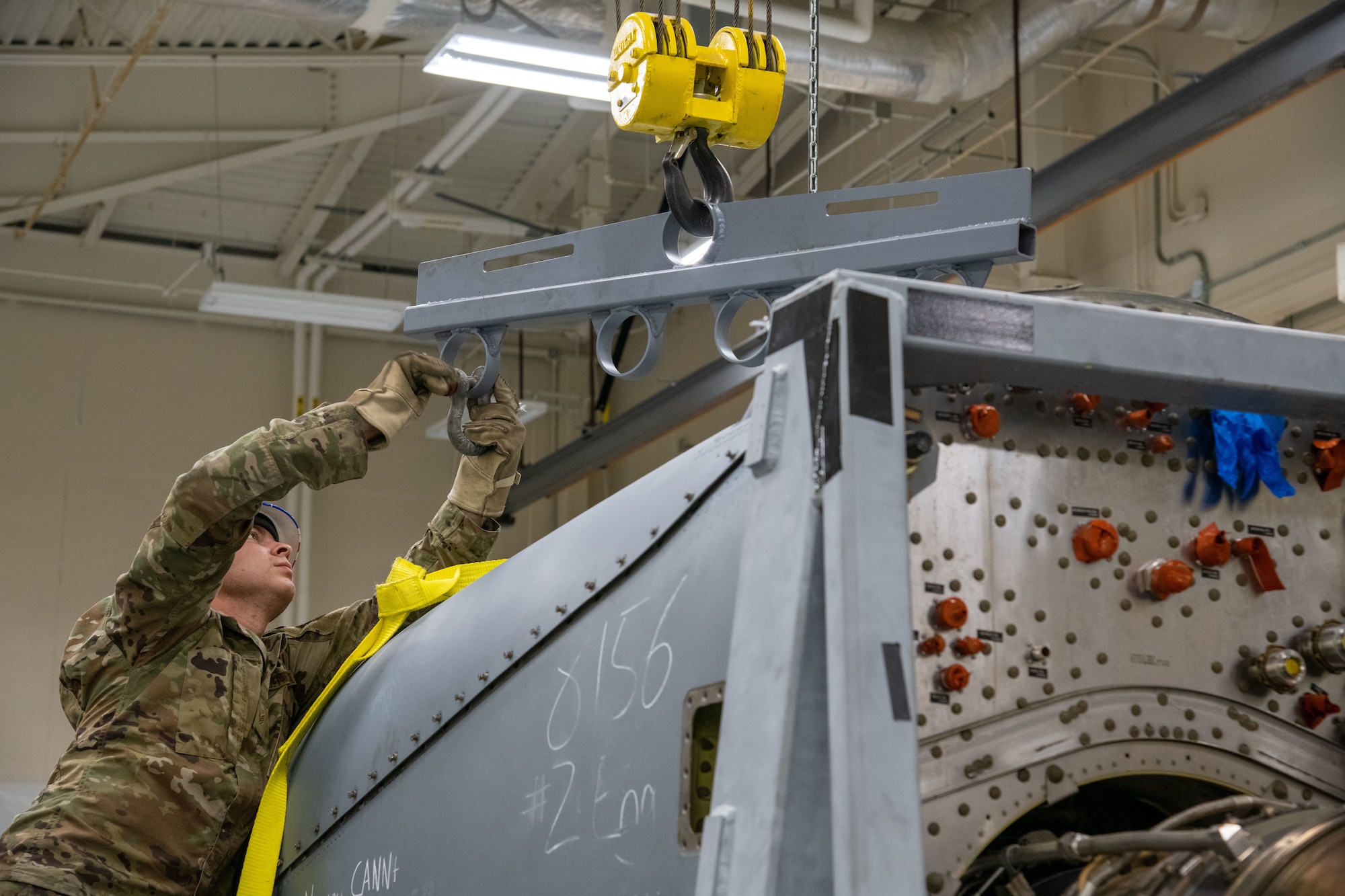 An Airman reaches above an engine to a metal piece hanging from a hoist to undo a shackle