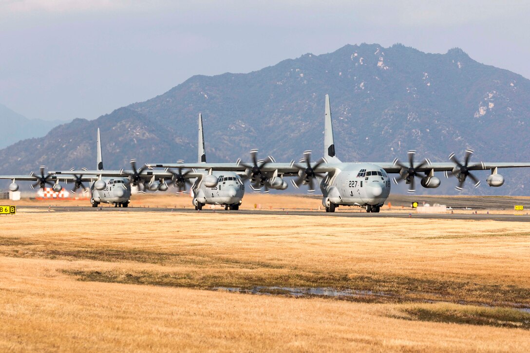 Three Marine Corps aircraft sit on a tarmac in a line in front of a mountain.