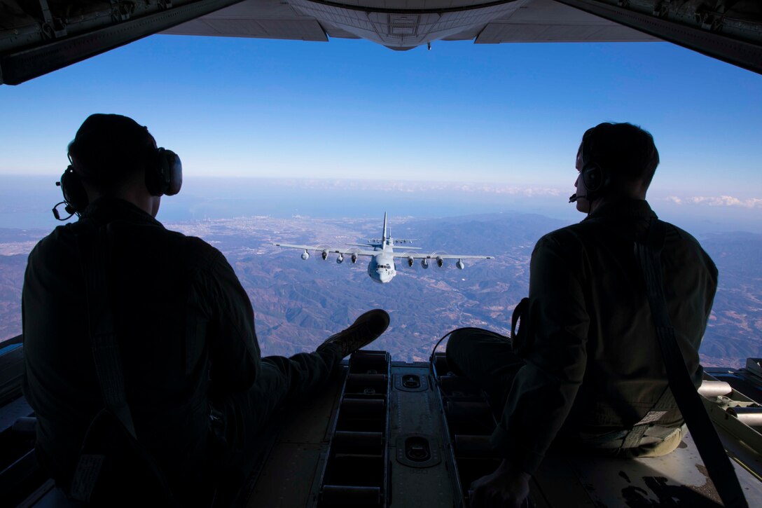 Two Marines sit at the back of an aircraft looking out at two airborne aircraft.