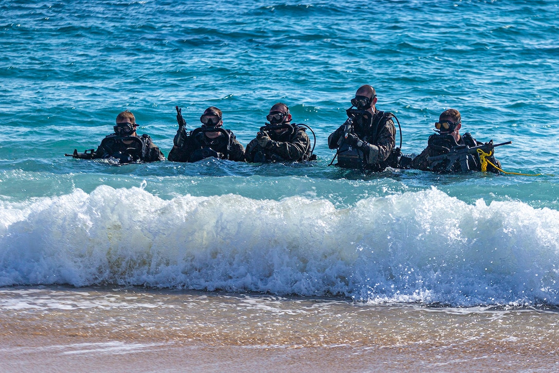 Five Marines walk through water toward a beach while wearing dive gear and holding weapons.